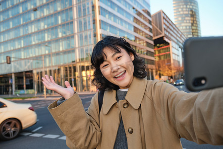 微笑的韩国女孩展示城市，在街上的建筑物前用快乐的脸自拍，用她的智能手机相机拍照