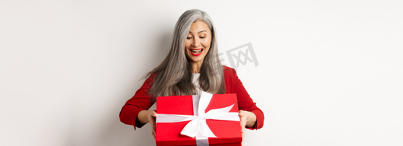 头发灰白的快乐老妇人收到礼物，看着红色礼盒，惊讶地笑着，站在白色背景上