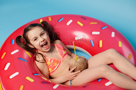 顶视图迷人的小女孩喝椰子饮料，在漂浮的甜甜圈游泳圈上放松，孤立的蓝色背景