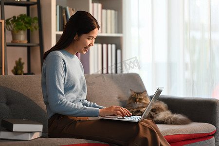 漂亮的亚洲女人在沙发上使用笔记本电脑，与家猫微笑。