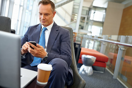 给他的客户发短信以确认会议......英俊的成熟商人坐在公司休息室的笔记本电脑前使用他的手机。