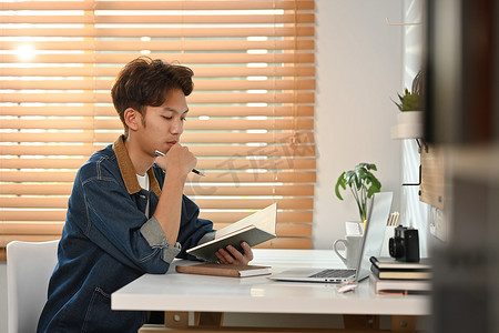 专注于牛仔裤夹克的亚洲男性自由职业者检查他的工作日程计划并在家庭办公室使用笔记本电脑