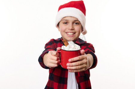 热男孩摄影照片_细节：一杯热巧克力加棉花糖，在戴着圣诞帽的模糊开朗男孩的手中，对着镜头微笑
