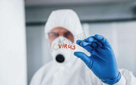 病毒防御摄影照片_身着实验室外套、防御眼镜和面具的男医生科学家拿着玻璃，上面有病毒词