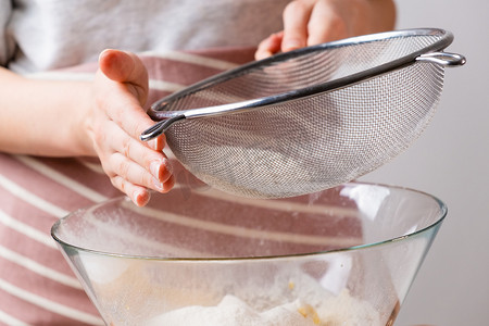分割线黑色过渡摄影照片_特写女人通过筛子将小麦粉筛入混合碗中，为黑色背景的蛋糕和面包店准备面团。