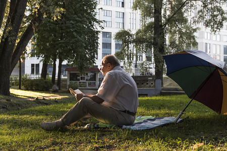 一位身穿白衬衫的老人坐在公园地上的毯子上，正在读一本有趣的书。