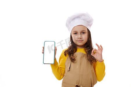 小女孩，厨师糖果师，用 OK 手势做手势，并在镜头前展示一部带有空白白色数字屏幕的智能手机