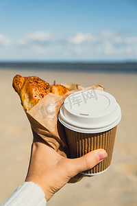 咖啡原料摄影照片_美味的新鲜羊角面包，在海滩大海背景下，女性手中的纸杯中装有咖啡。