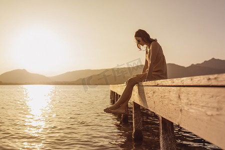 温情情绪摄影照片_沉思的单身白人漂亮年轻女子坐在湖边的码头上，在日落时俯视水面 — 复古橙色的心情