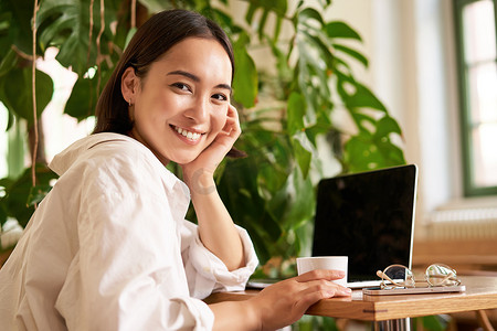 时髦的年轻女性坐在咖啡馆里，对着镜头微笑、喝咖啡、使用笔记本电脑、远程工作、学习或浏览互联网