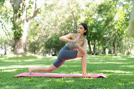 年轻迷人的女孩正在公园中央的健身垫上做高级瑜伽体式。