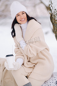 一个穿着米色开衫和白帽子的漂亮女孩在湖边的雪冬森林里喝茶