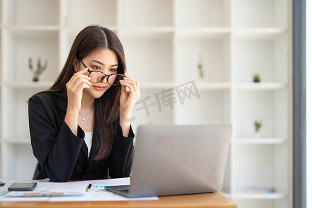 金融美女摄影照片_照片中一位美女戴着眼镜坐在办公桌前，周围是办公室的笔记本电脑、文书工作、计算器和咖啡杯。