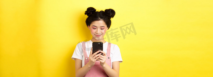 时尚的亚洲女孩，用手机化着迷人的妆容，对着屏幕微笑，站在黄色背景上