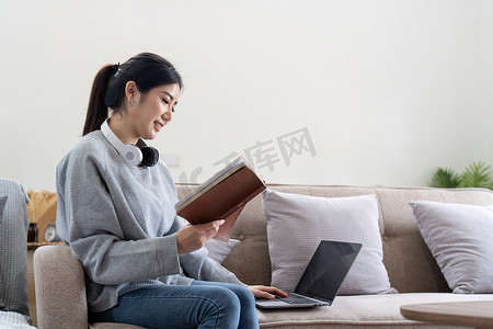 火爆进行中摄影照片_年轻漂亮的亚洲女学生坐在沙发上在家看书，同时使用笔记本电脑进行在线学习