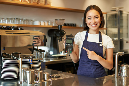微笑的亚洲女咖啡师，围裙的咖啡馆老板，展示卡片机，支付读卡器，在她的咖啡店接受非接触式订单