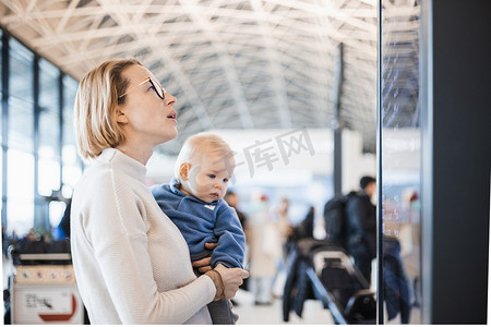 母亲抱着孩子摄影照片_母亲带着孩子旅行，在机场航站楼抱着他的婴儿男婴，查看航班时刻表，等待登机。