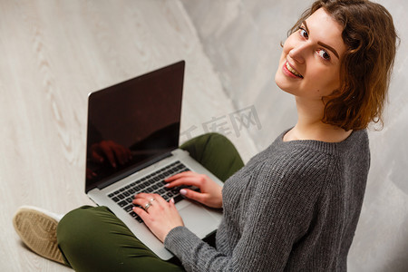 满意的女性画像，带着美丽的微笑，享受着用银色电脑看电影，坐在灰色墙壁上的地板上的莲花姿势