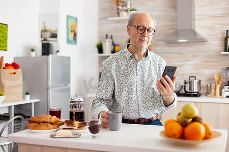 老人技术摄影照片_快乐的老人在早餐时使用智能手机在社交媒体上冲浪