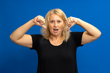 40 多岁的金发白人女性在蓝色工作室背景下厌恶地竖起大拇指。