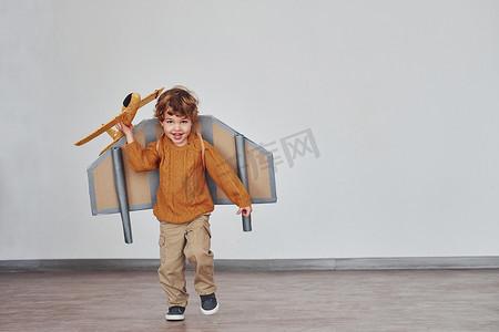材料员摄影照片_穿着复古飞行员制服的小男孩在室内带着玩具飞机跑