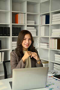 亚洲女经理坐在办公桌笔记本电脑前，对着镜头微笑的画像