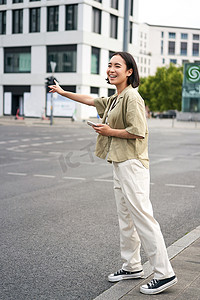 共享汽车摄影照片_垂直拍摄的年轻女性试图赶出租车，在路上向司机挥手，手持带有汽车共享应用程序的智能手机，站在城市街道上