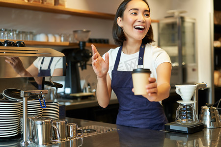 微笑的亚洲女咖啡师的画像，在咖啡厅下订单，邀请客人在柜台附近取外卖订单，拿着外卖咖啡