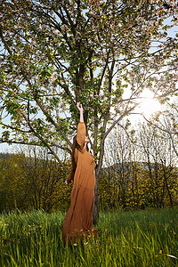 一位长着红色长发的苗条女性穿着橙色长裙站在一棵开花的树旁，侧身站在镜头前伸手去拿树枝