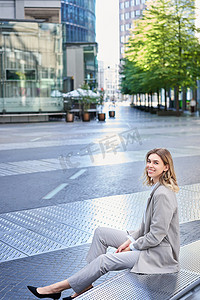 公司大楼照片摄影照片_身着米色西装、坐在商务办公大楼外、自信地看着的微笑女商人的垂直照片