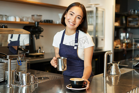 漂亮的年轻女咖啡师，做卡布奇诺，把拿铁艺术的蒸牛奶倒进杯子里，在柜台后面下单