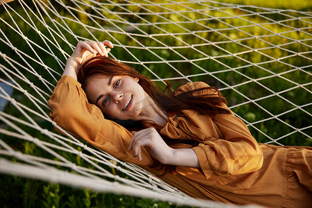 一个快乐的女人躺在网眼吊床上休息，头枕在手上，对着镜头开心地笑着，穿着橙色的连衣裙，在夕阳的余晖中享受着温暖的一天