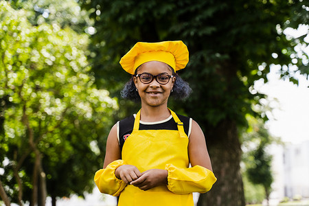黑人厨师摄影照片_穿着厨师帽和黄色围裙制服的黑人非洲儿童厨师女孩在户外微笑。