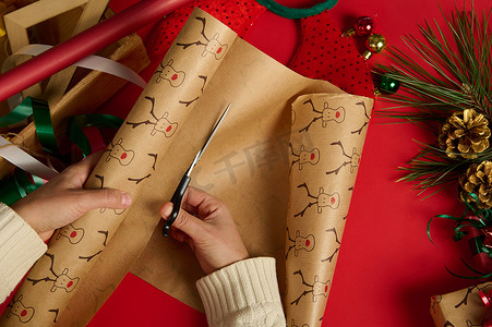 穿着米色毛衣的顶视图女人用剪刀，在红色背景上剪下包装礼品纸。