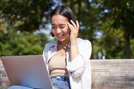 美丽的亚洲女孩坐在公园的长椅上笑着，在笔记本电脑上看视频，听音乐或在线视频聊天