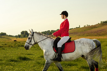 年轻的女骑手，穿着红色的红色长裤和白色的马裤，在夕阳的余晖中骑着她的马。