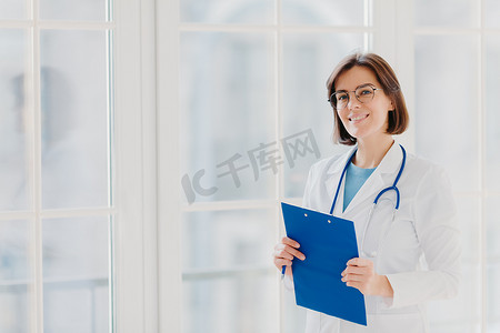 女性心脏治疗师戴圆眼镜、白色医用外套和语音内窥镜，用笔在剪贴板上写字，站在自己诊所的大窗户附近，思考病人的病例和治疗