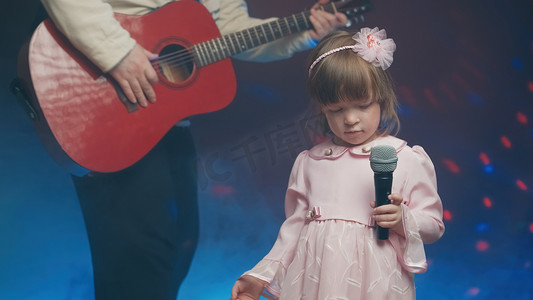 儿童节音乐摄影照片_穿着复古裙子的小女孩在舞台上唱歌，她的父亲弹着原声吉他