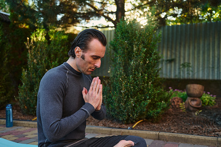 特写一个男瑜伽士，在健身垫上以莲花姿势冥想，双手合十，在日落时练习瑜伽