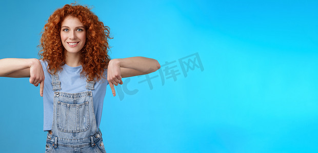 魅力四射的时髦红头发大胆的姜黄色女孩卷发指着食指微笑着广泛热情地探索新店指向促销就像酷广告蓝色背景