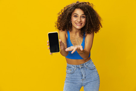 手持电话的有影响力的女性在视频通话中自拍自由职业者社交媒体技术工作，蓝色衬衫黄色背景的卷发爆炸头，牙齿微笑，复制空间