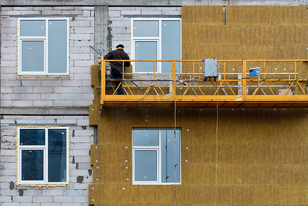 外墙施工摄影照片_一名建筑工人站在悬空的平台上，用矿物隔热材料对在建高层建筑的外墙进行隔热。