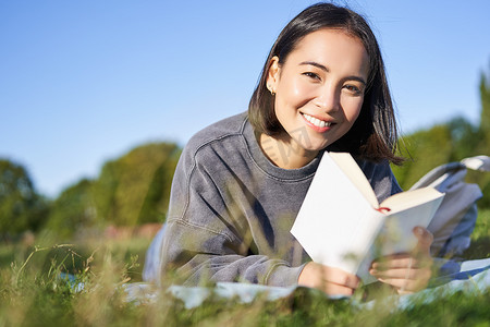 可爱的韩国女孩的画像，在公园里躺在草地上读书，手里拿着最喜欢的书放松，开心地笑着