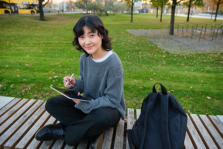 带着图形铅笔和平板电脑的亚洲小女孩坐在公园的长椅上，画画，在户外做作业