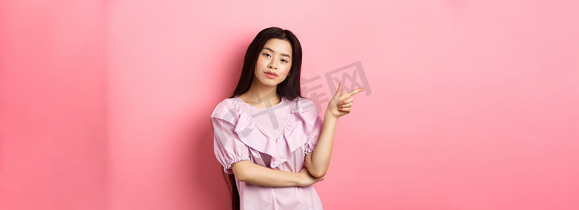 年轻自鸣得意的亚洲女孩看起来很酷，手指留在标志上，在粉红色浪漫背景下做广告产品