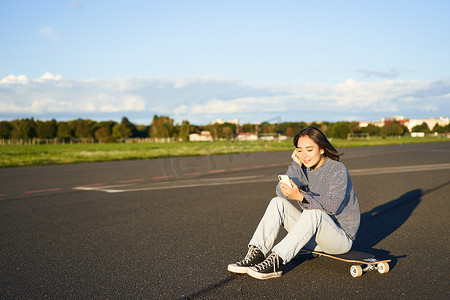 溜冰女孩坐在路上的滑板上，使用智能手机，在移动应用程序上聊天