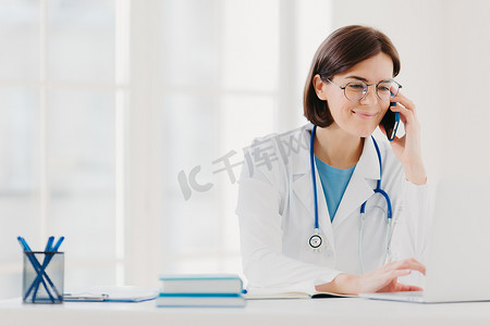 微笑的治疗师专注于笔记本电脑屏幕，通过现代智能手机打电话，穿着医疗长袍，戴着透明眼镜，坐在联合办公空间，上网冲浪。