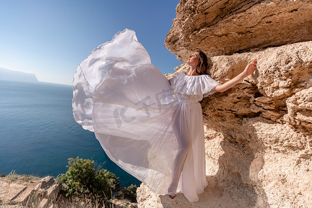 在蓝天大海的映衬下，一位身穿白色轻裙长腿的美丽少妇挥舞着白色长裙站在海面之上的悬崖边上。