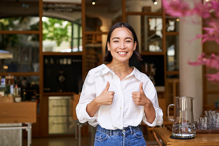 快乐的亚洲女人竖起大拇指，赞成，喜欢好东西，站在咖啡馆入口附近，推荐她