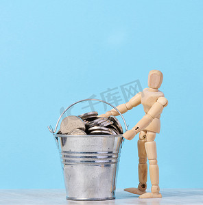 补贴摄影照片_蓝色背景中微型桶中的木制人体模型和硬币，高收入、补贴的概念